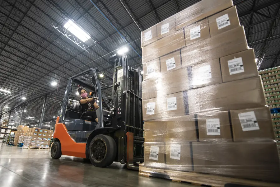 Forklift operator moving stacked boxes in a large warehouse